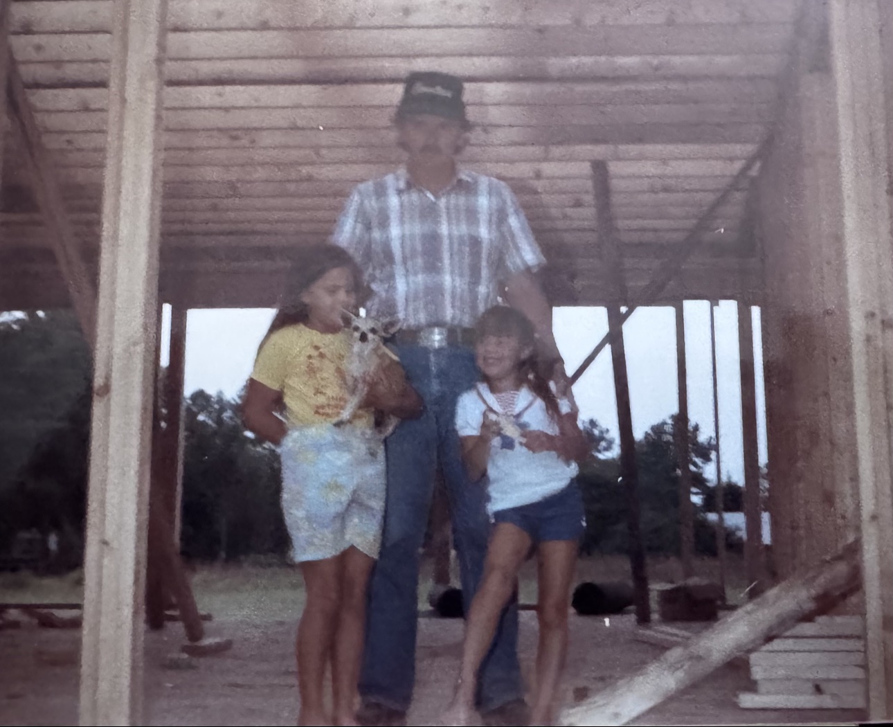 Tiffany McGhee's dad standing with a young Tiffany and her sister Stephanie, holding a little dog, inside the wooden framing of the first house he ever built