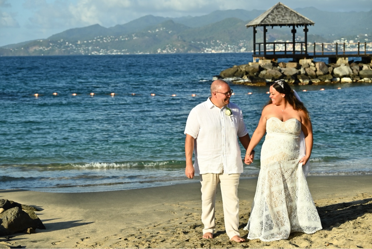 Tiffany McGhee in her wedding gown walking hand in hand with her husband Michael along the beach in Grenada, with a gazebo pier and mountains behind them